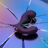 A Svakom Iker App-Controlled Prostate and Perineum Vibrator sits on a reflective, cracked surface with a colorful gradient. The cracks radiate from the center, creating a dynamic pattern beneath the sleek, black vibrator.